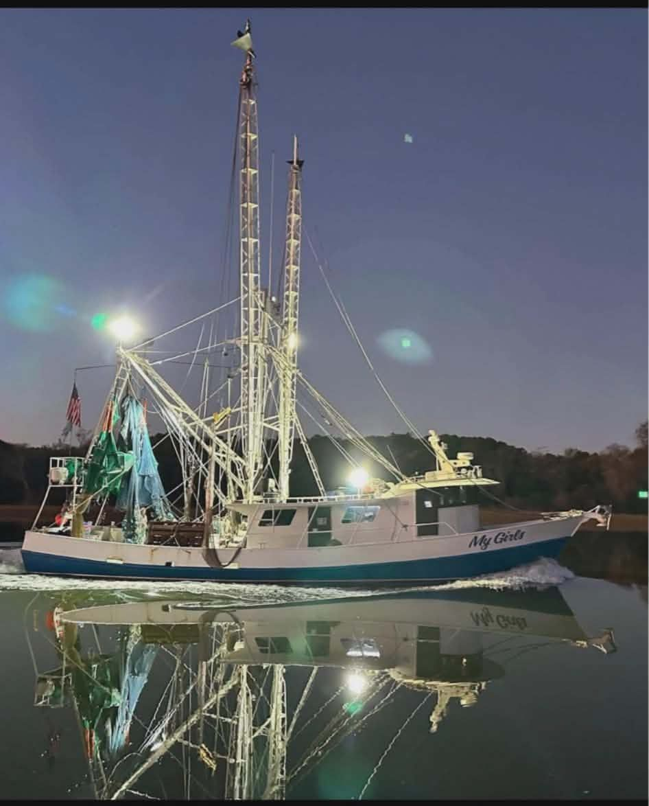 Fishing boat sailing at night with illuminated rigging, reflected in the water.