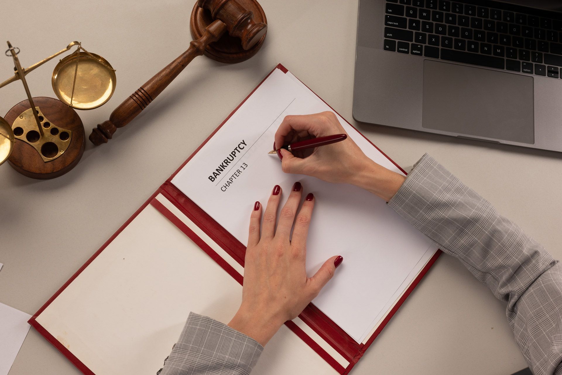 A person in a gray patterned blazer writes on a document in a red folder, beside a wooden gavel and scales of justice.