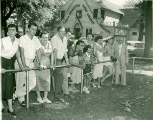 Group of people leaning on a railing outdoors, observing something. Building with sign in the background.