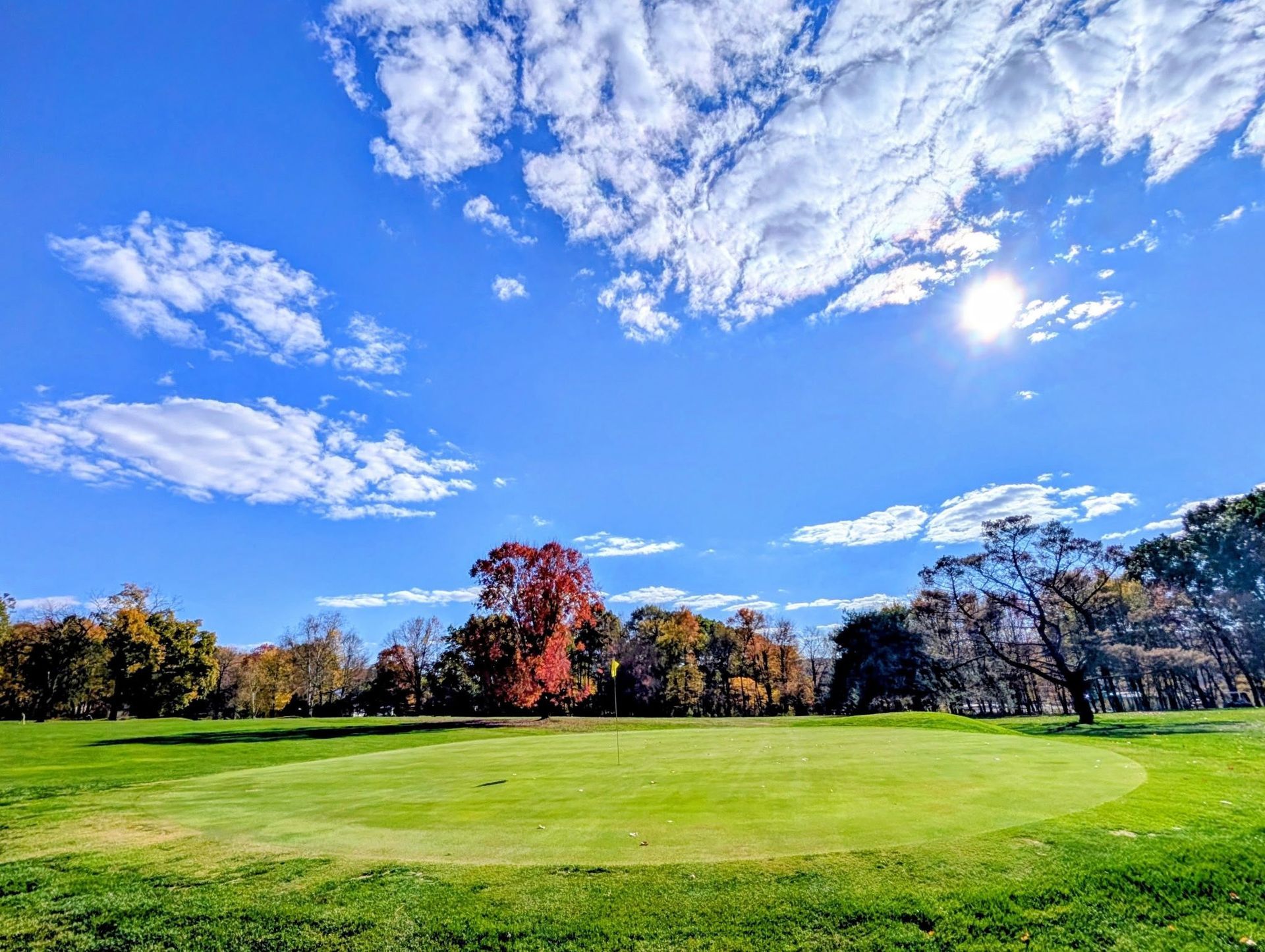 Golf course green with red and yellow trees under a bright blue sky with clouds.