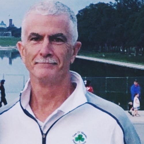 Man with gray hair and mustache in a white jacket, posing near a body of water in a park.