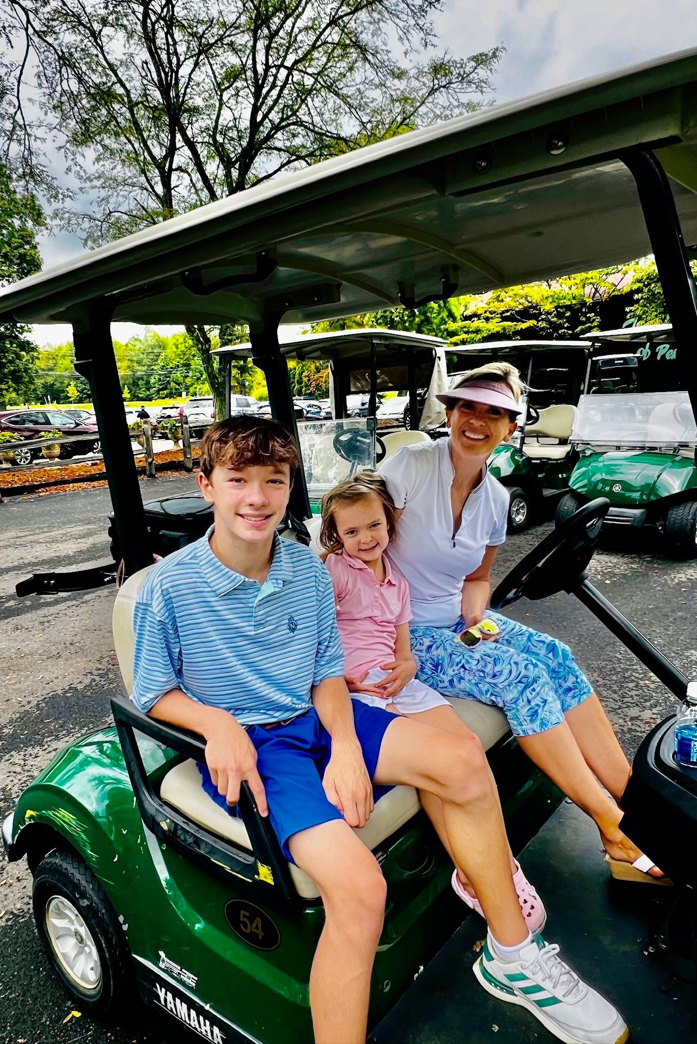 Family of three in a green golf cart, smiling. Outside in a parking lot, sunny day.