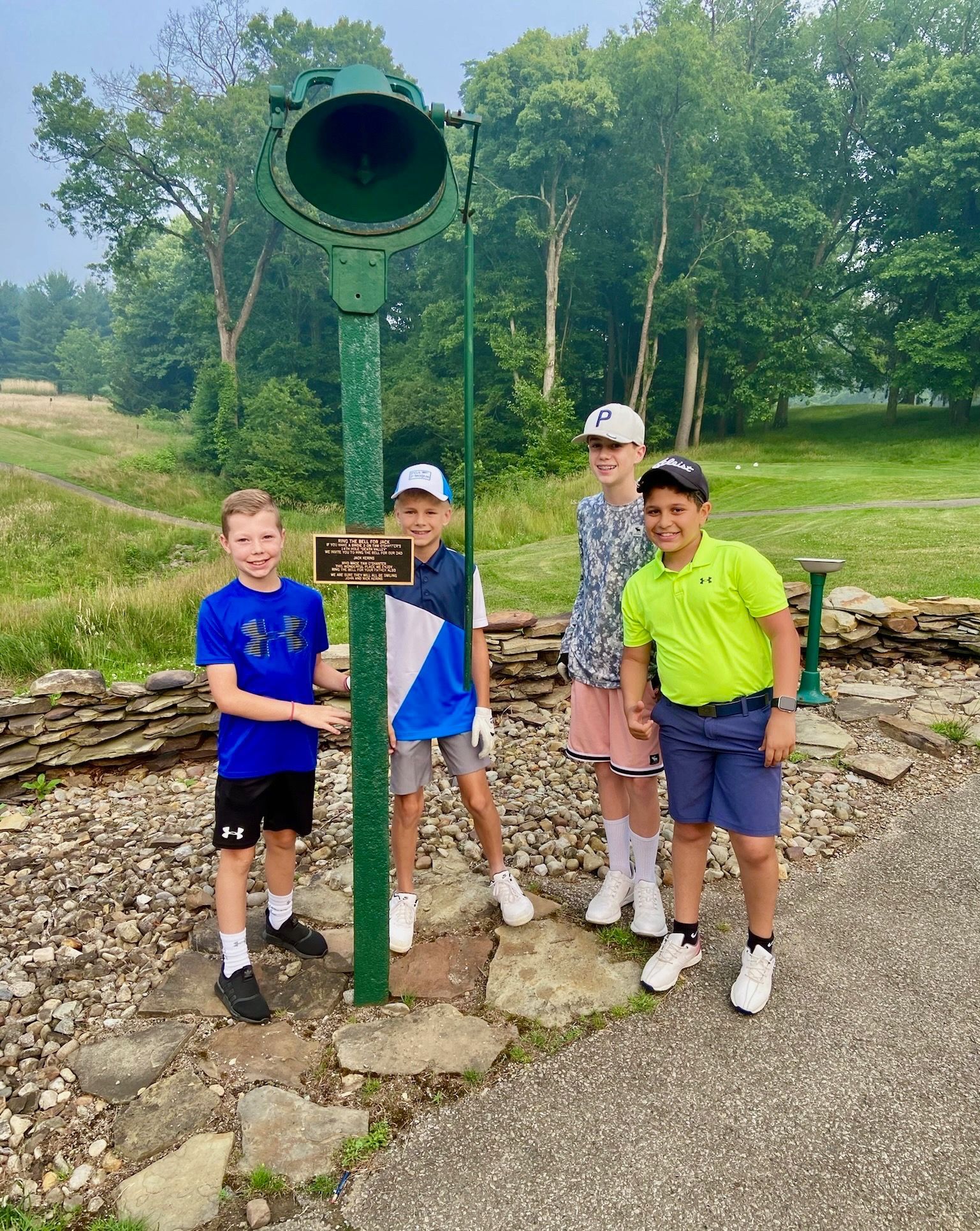 Four boys stand near a green bell on a golf course, smiling at the camera on a sunny day.