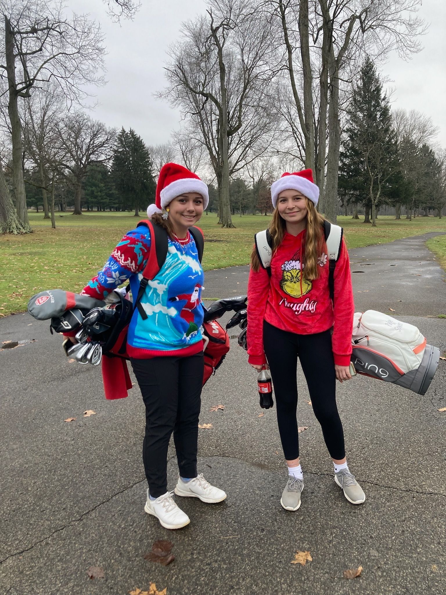 Two people in Santa hats and sweaters on a golf course. One carries clubs, the other holds a soda.