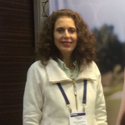 Woman in a white jacket with curly brown hair, wearing a badge, smiling, indoors.