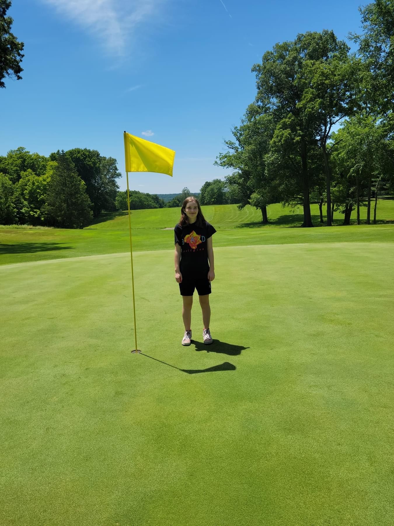 Person stands on a golf green near the hole, yellow flag in the background, blue sky and trees.