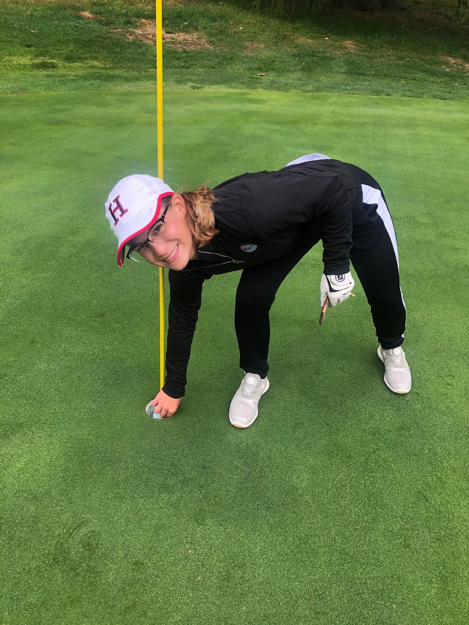 Golfer on green, leaning to pick up ball near a yellow flag, wearing white hat and shoes.