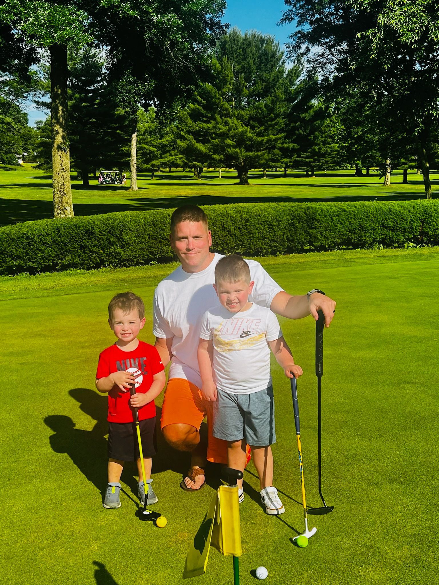Man and two young children on a golf course holding putters. Green grass and trees in the background.