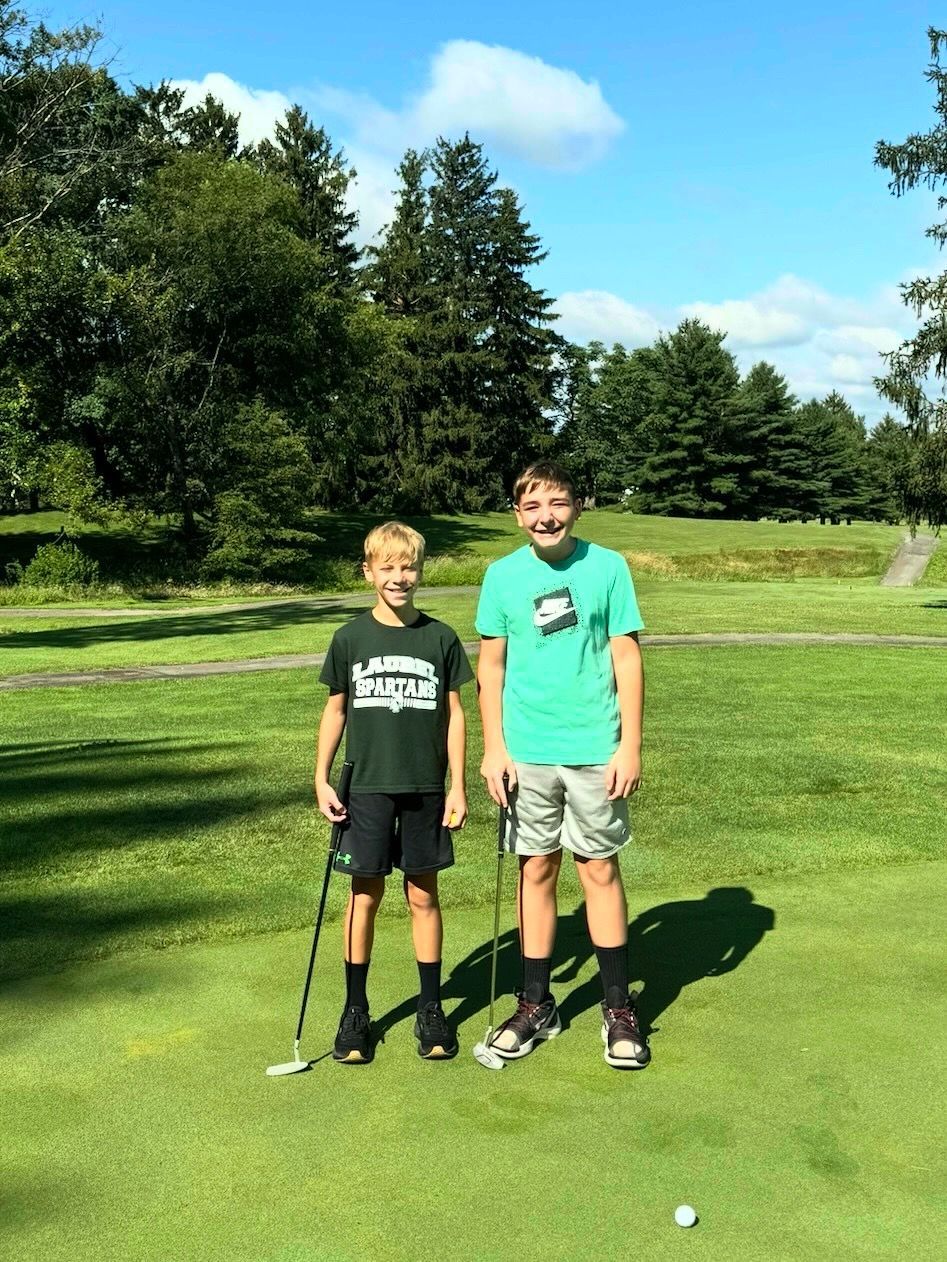 Two boys standing on a golf course green, holding golf clubs. Bright, sunny day.