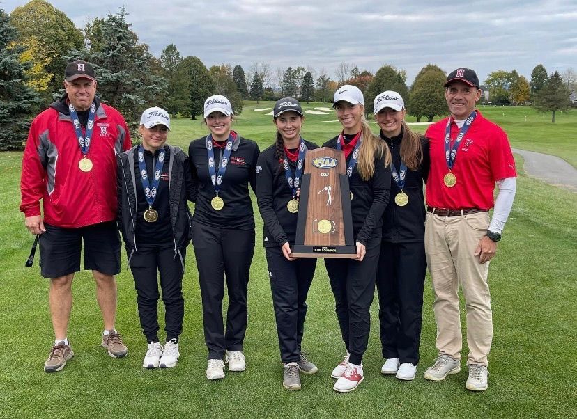 Golf team with trophy and medals on a green golf course, wearing black, red and white attire.