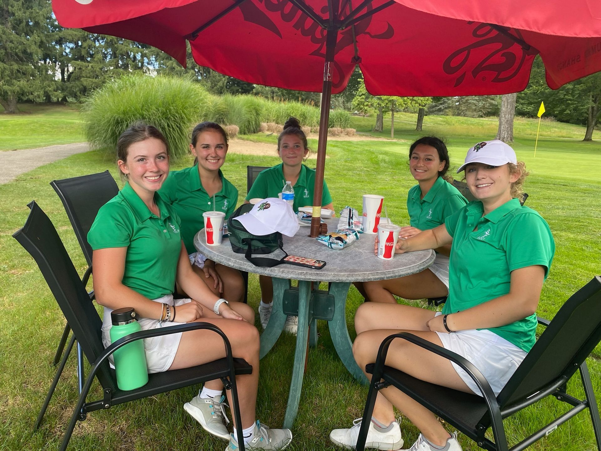 Golf team, wearing green shirts and white shorts, sitting under an umbrella at a table on the course.