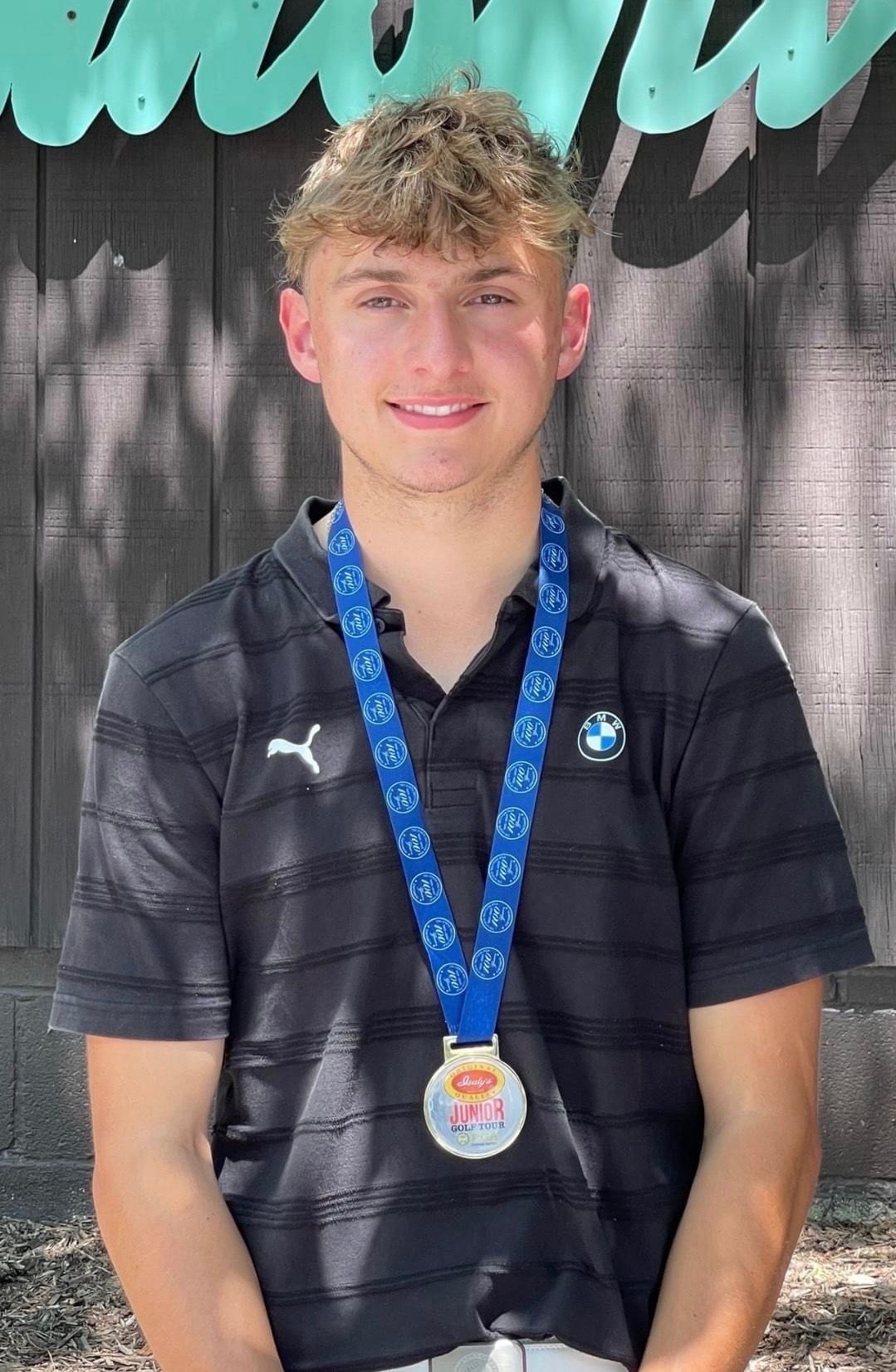 Young person wearing a medal, smiling. Standing in front of a wooden backdrop with a blue logo.