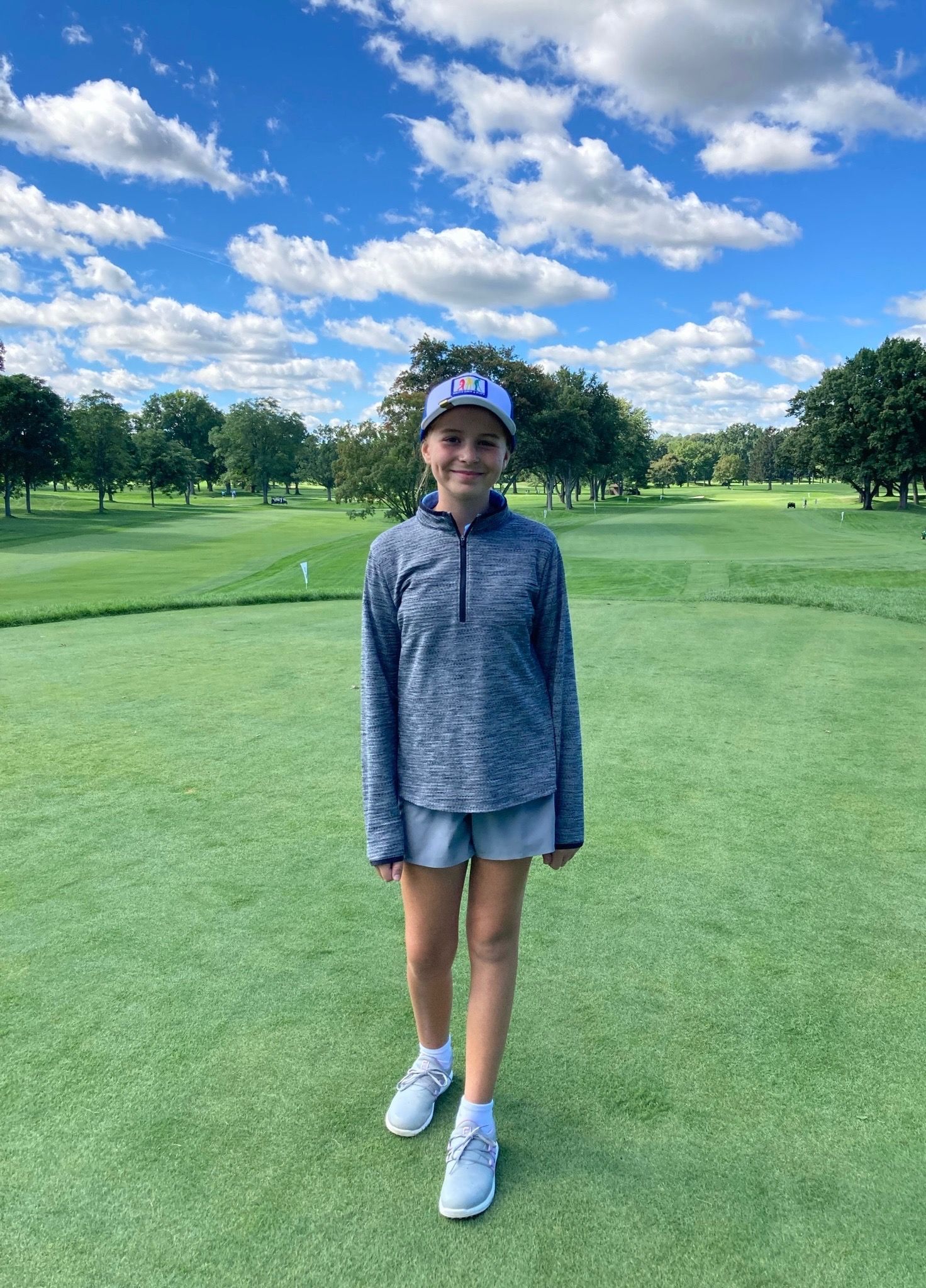 Young person in golf attire on a golf course under a blue sky with fluffy clouds.