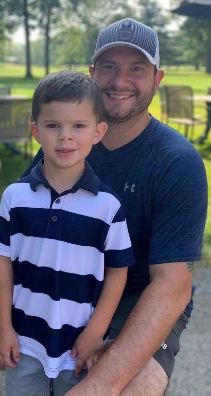 Man holds a young child outdoors; both smiling. The child wears a striped shirt, man in a hat and blue shirt.