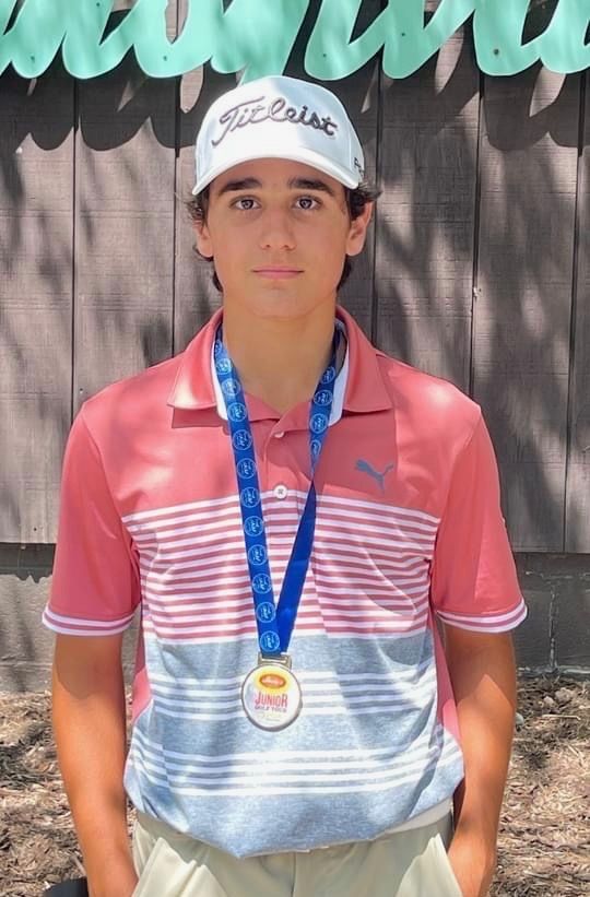 Young golfer wearing a medal, Titleist hat, and pink polo shirt, standing in front of a wooden backdrop.