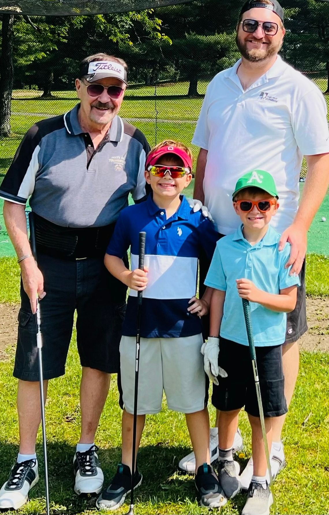 Four people on a green golfing range. Two young boys in sunglasses hold clubs, flanking two adults, who also have clubs.