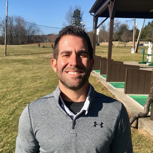 Man smiling at golf range, wearing a gray pullover.