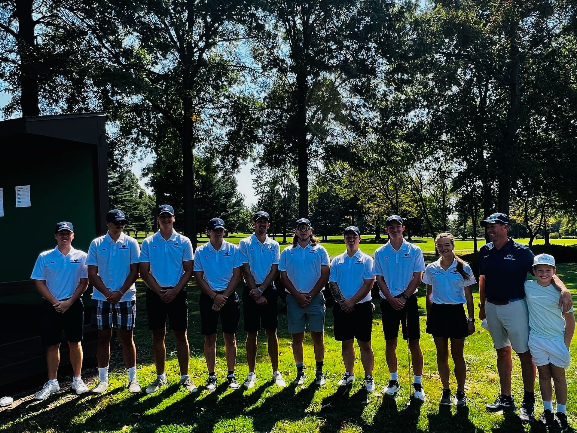 Golf team posing outdoors, wearing white shirts and dark shorts, on a sunny day near trees and a green structure.