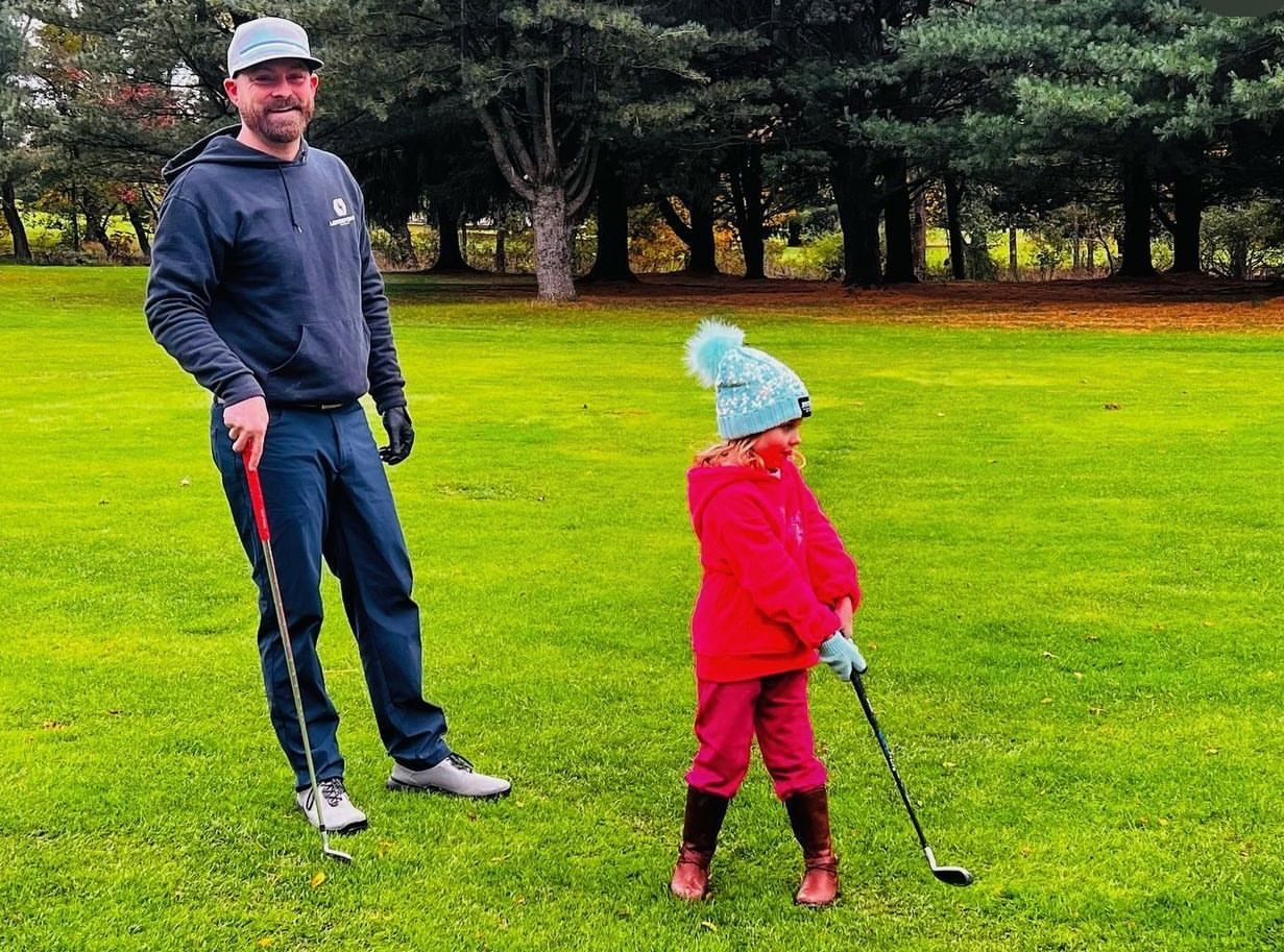 Man watches a child swing a golf club on a green lawn. They wear warm clothing, and trees are in the background.