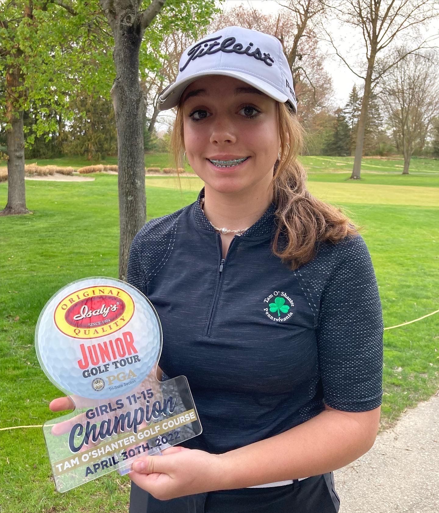 Girl with braces and Titleist cap holds golf trophy on course.