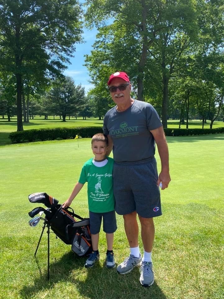 Man and boy on a golf course posing together, boy with golf bag, both smiling, green grass, sunny day.