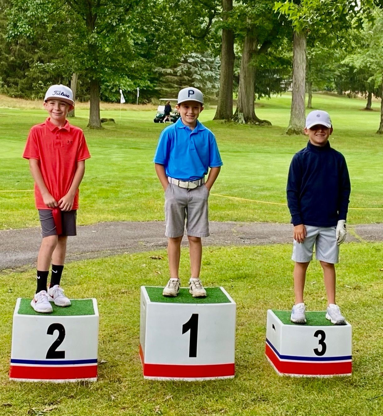 Three young golfers on a podium, numbered 1-3. Blue sky, green grass.