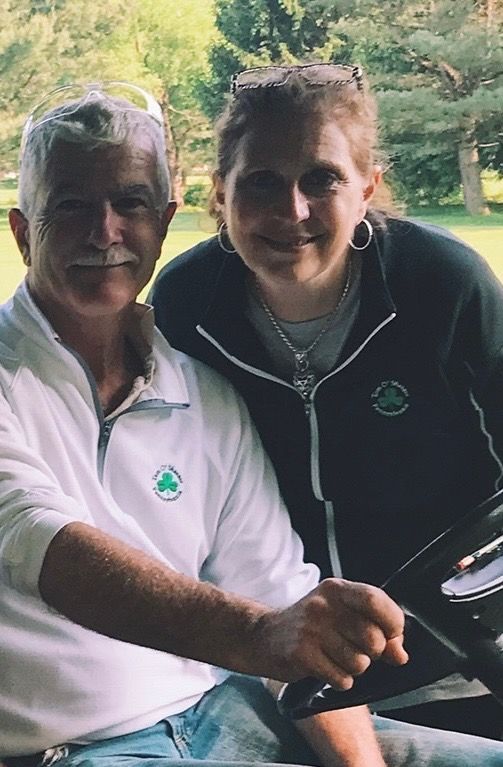 Man and woman smiling, seated in a golf cart. Man wears white golf shirt, woman wears a hoodie. Green, outdoor setting.