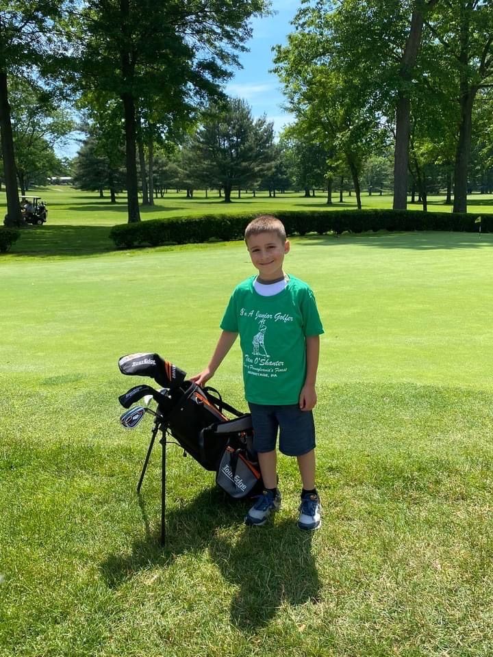 Young boy in a green shirt and shorts stands next to golf bag on a golf course.