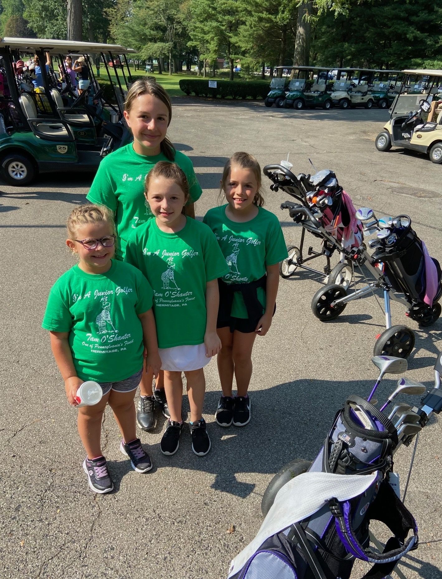 Four girls in green shirts at a golf course, posing near golf carts and bags.