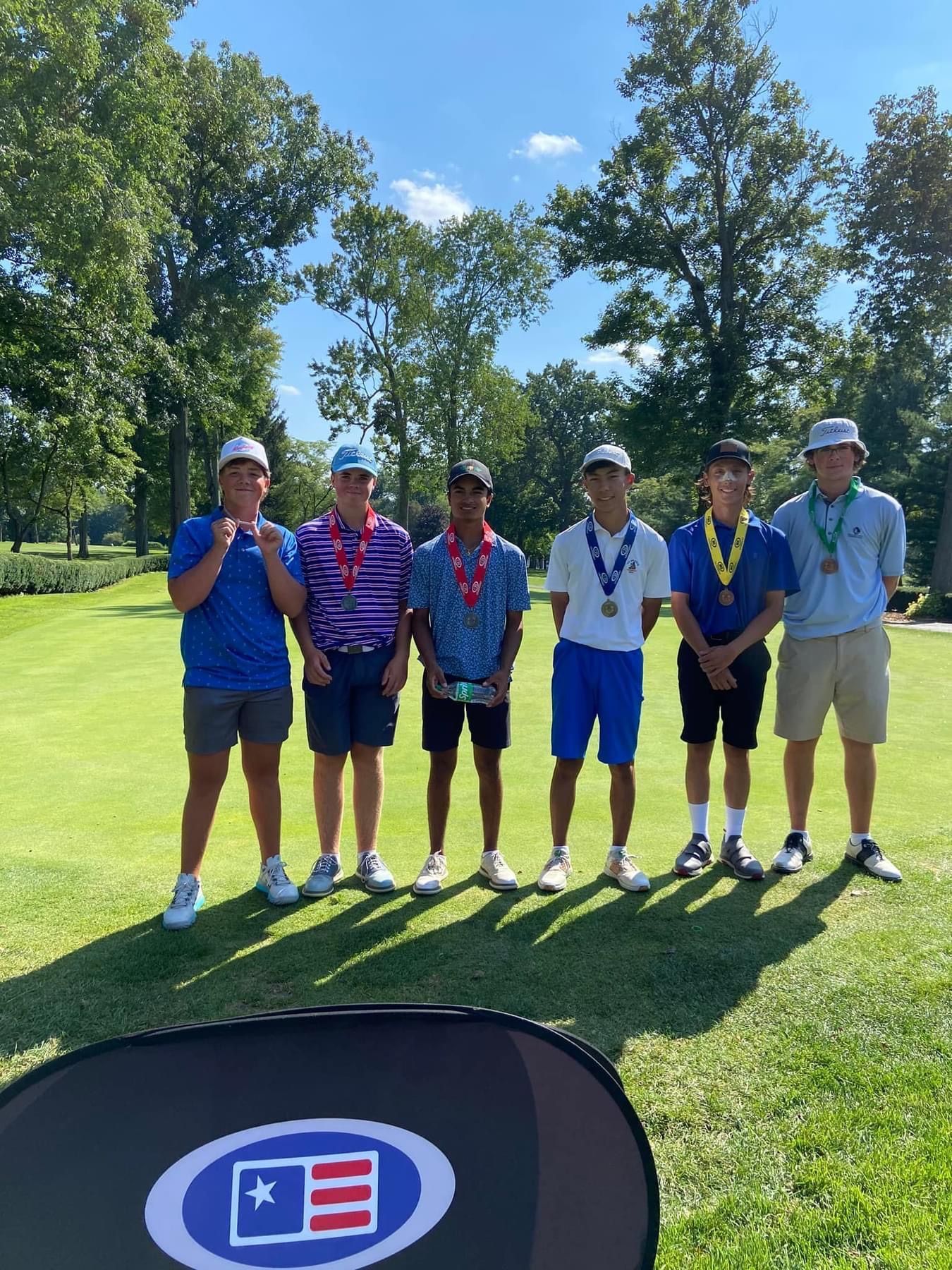 Group of six golfers wearing medals stand on a green, sunny golf course.