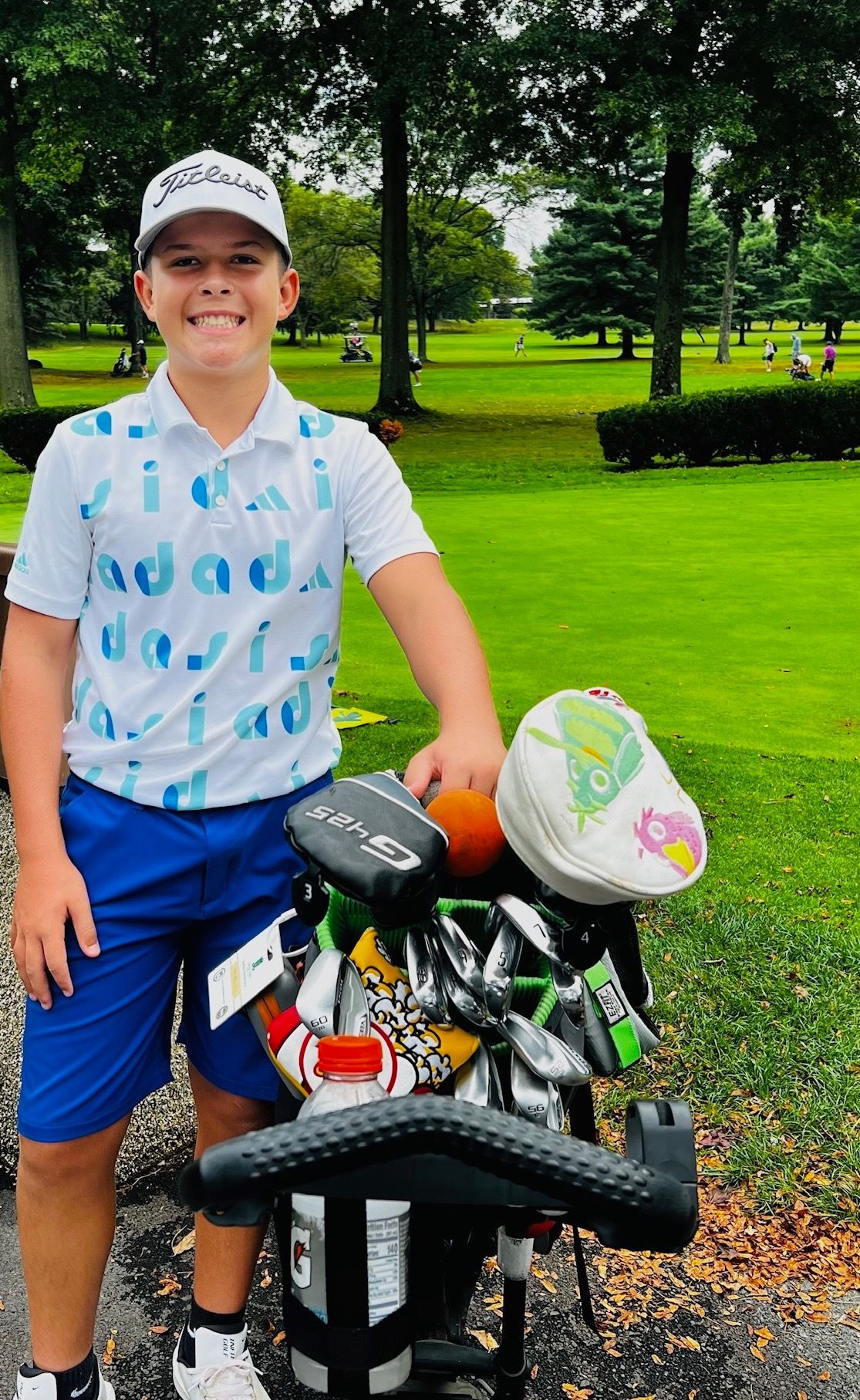 Young golfer smiles next to golf bag on green course, wearing blue shorts and a patterned shirt.