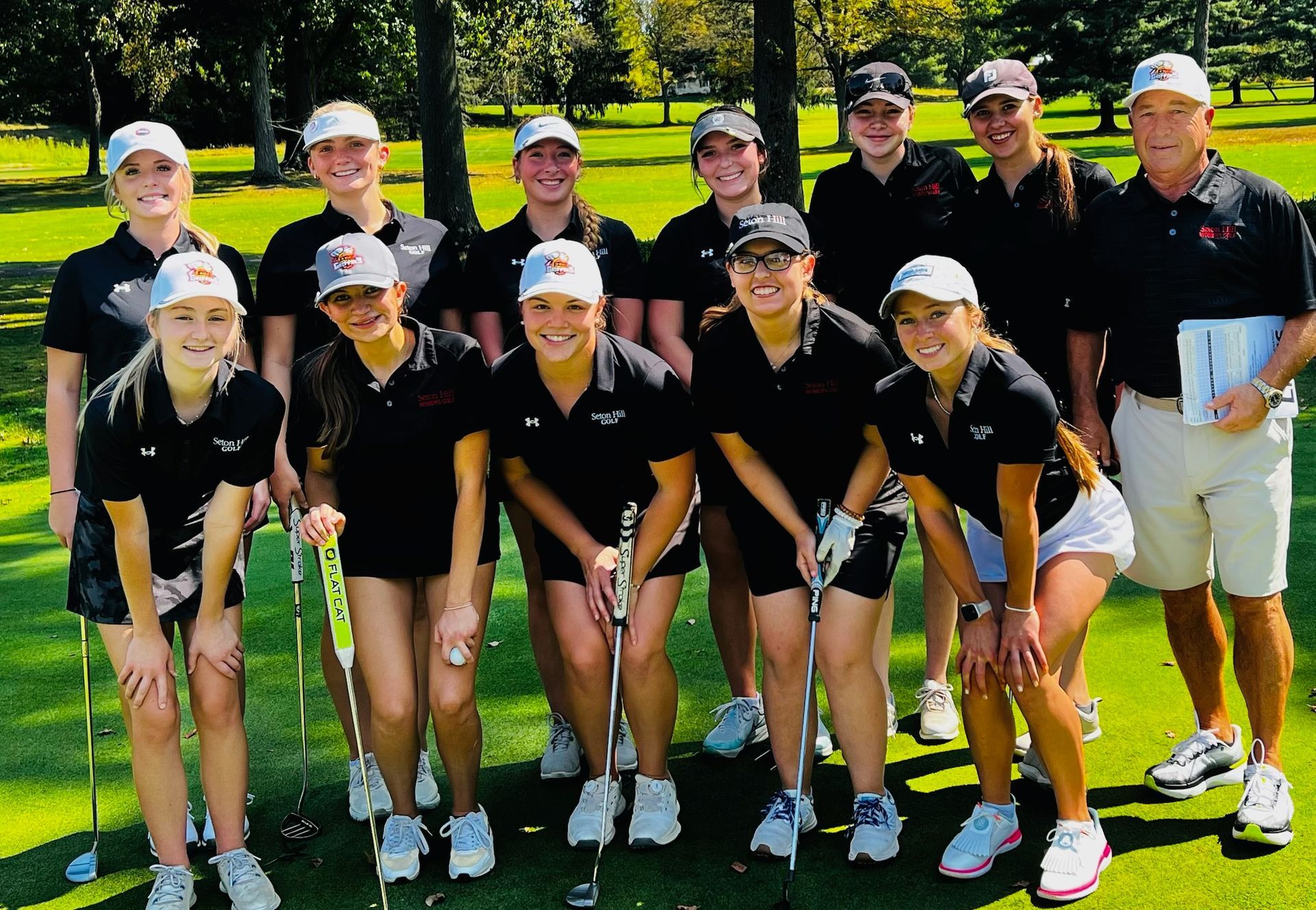 Group of golfers in black shirts and hats on a green, smiling with coach.