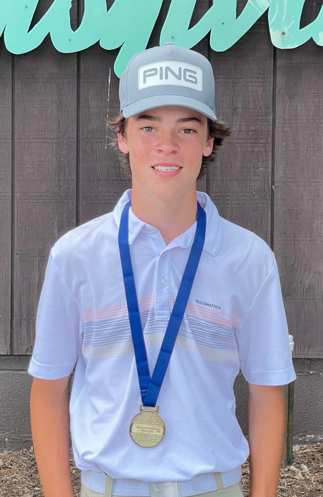Young person in a Ping hat wearing a medal, smiling. Standing in front of a wooden wall.