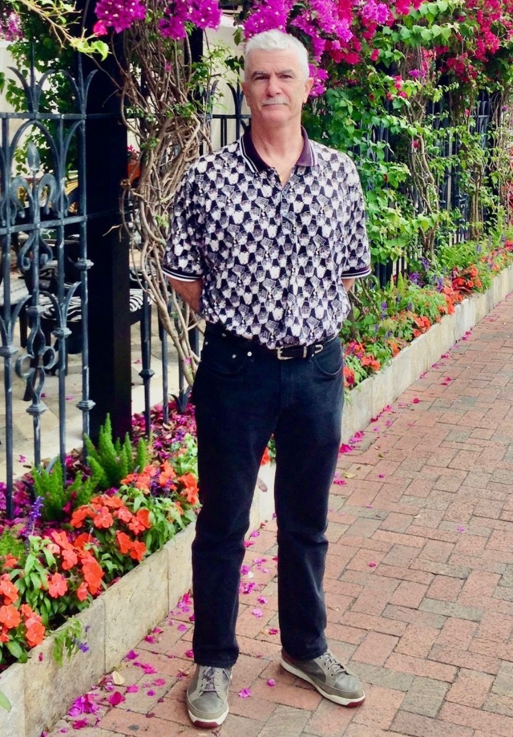 Man standing on a brick walkway, wearing a checkered shirt and dark jeans, with flowers and a wrought-iron fence behind him.