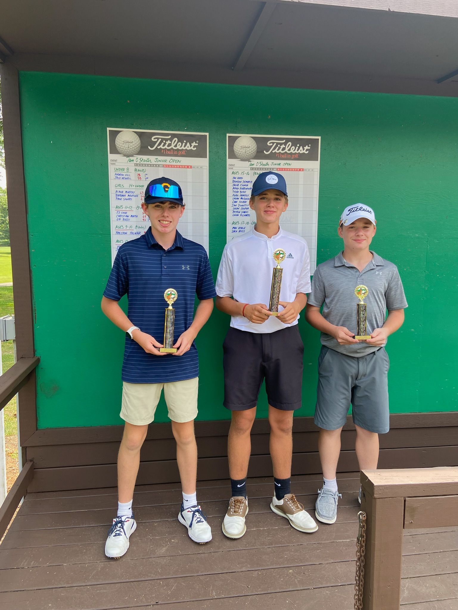 Three young golfers holding trophies in front of a green wall with scoreboards.