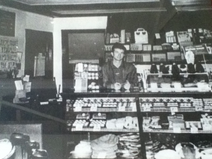 Man stands behind the counter of a small store, shelves stocked with various products. Black and white.