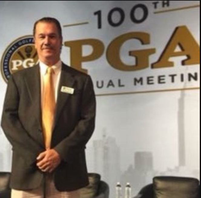 Man in suit stands before PGA Annual Meeting backdrop.