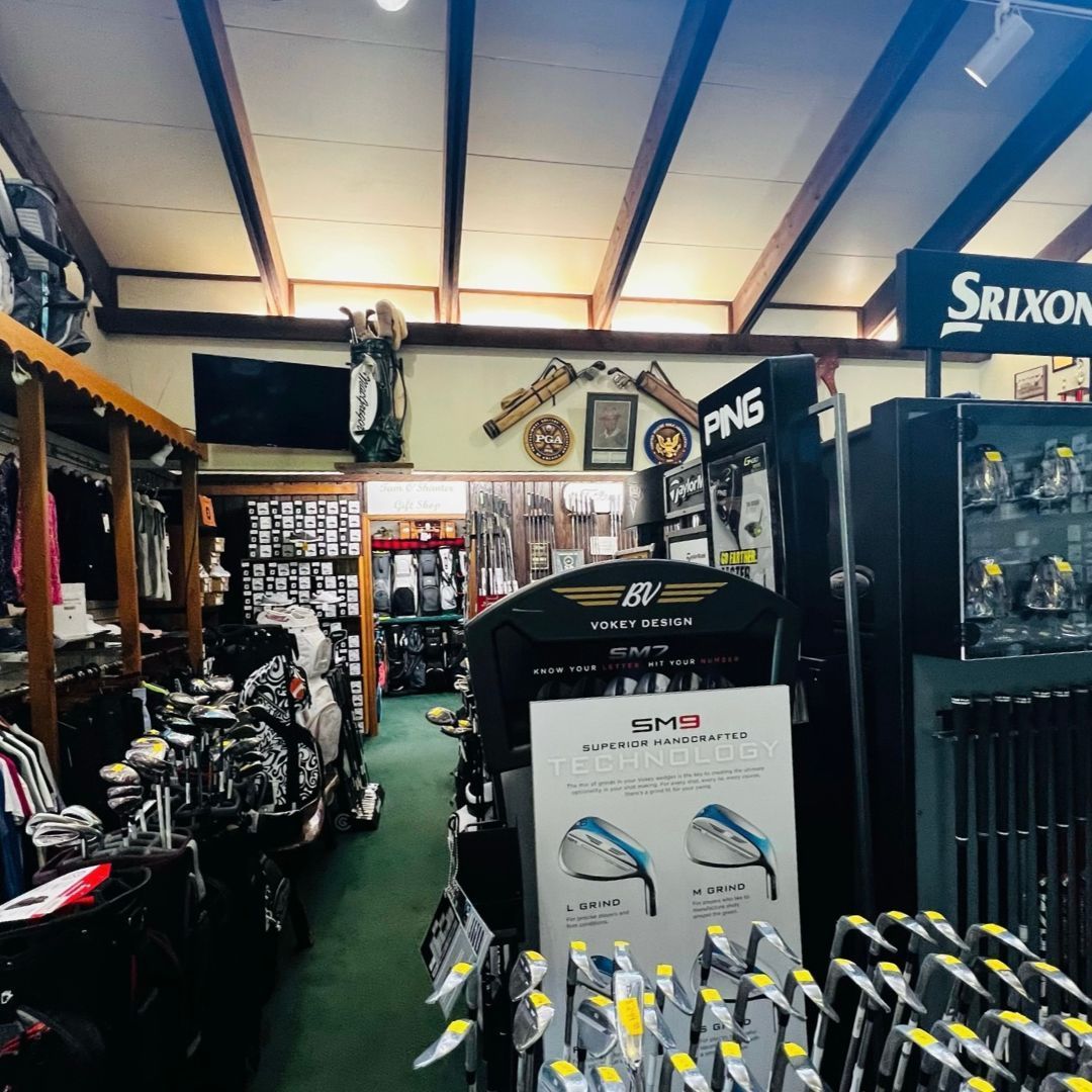 Golf shop interior with clubs, bags, and displays. Srixon and Ping signs visible. Wooden ceiling, green carpet.