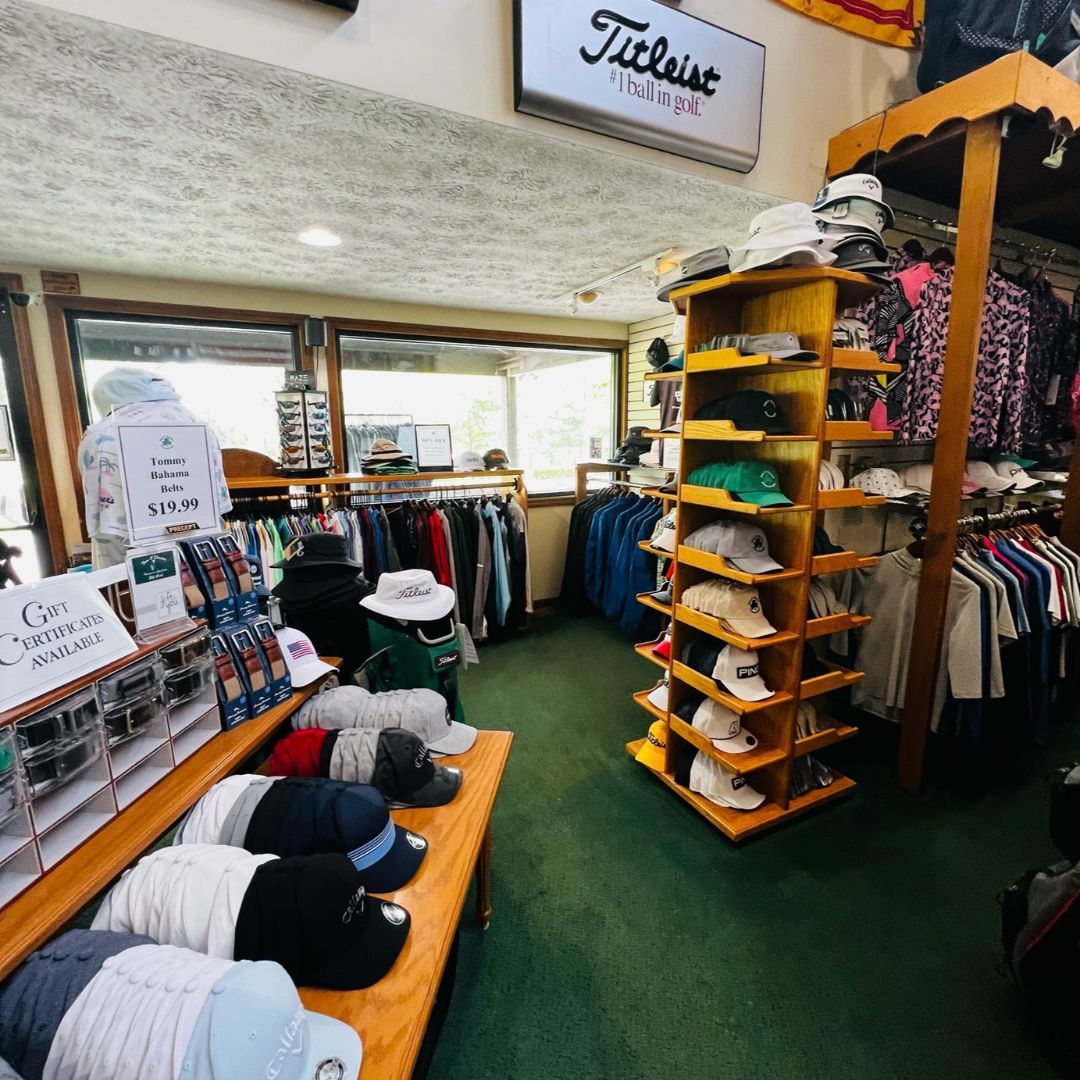 Golf shop interior with hats, apparel, and a Titleist sign. Green carpet, wooden shelves, natural light.