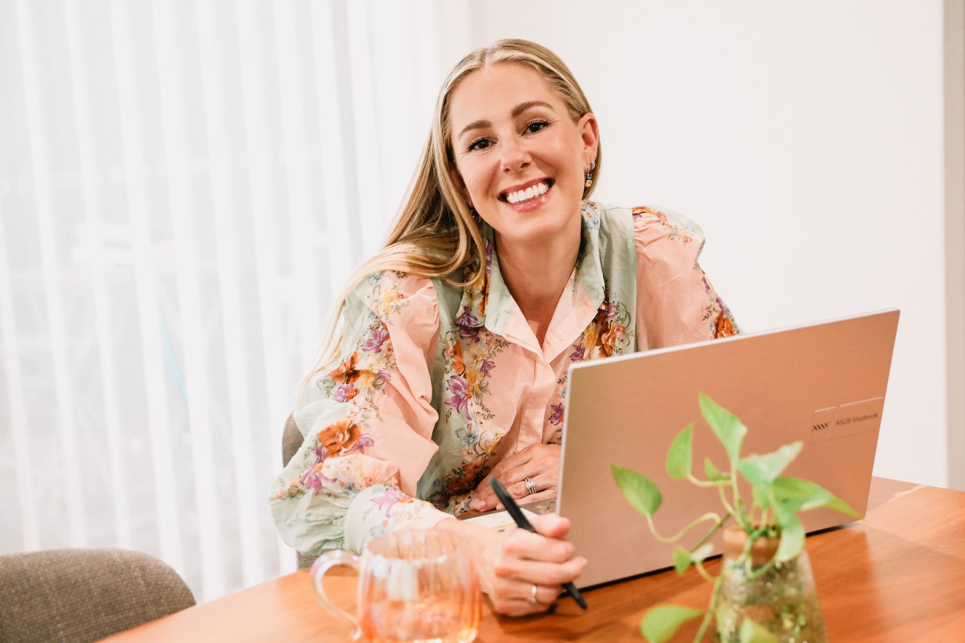 Woman smiling, working on laptop at a wooden table in a well-lit room, with a houseplant and cup.