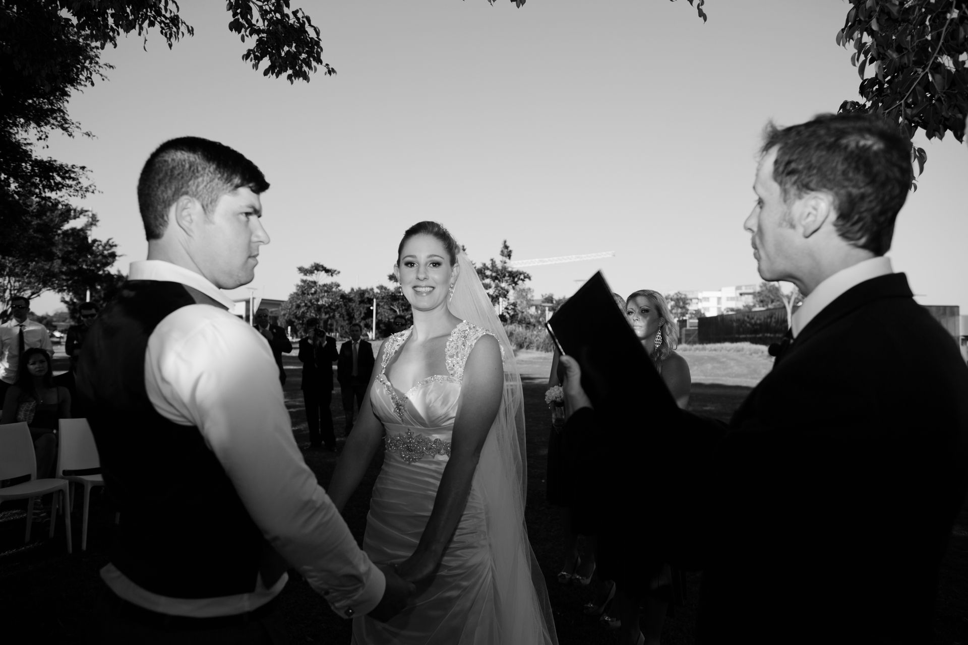 Wedding ceremony with a couple holding hands, facing a person with a book; outdoor setting.