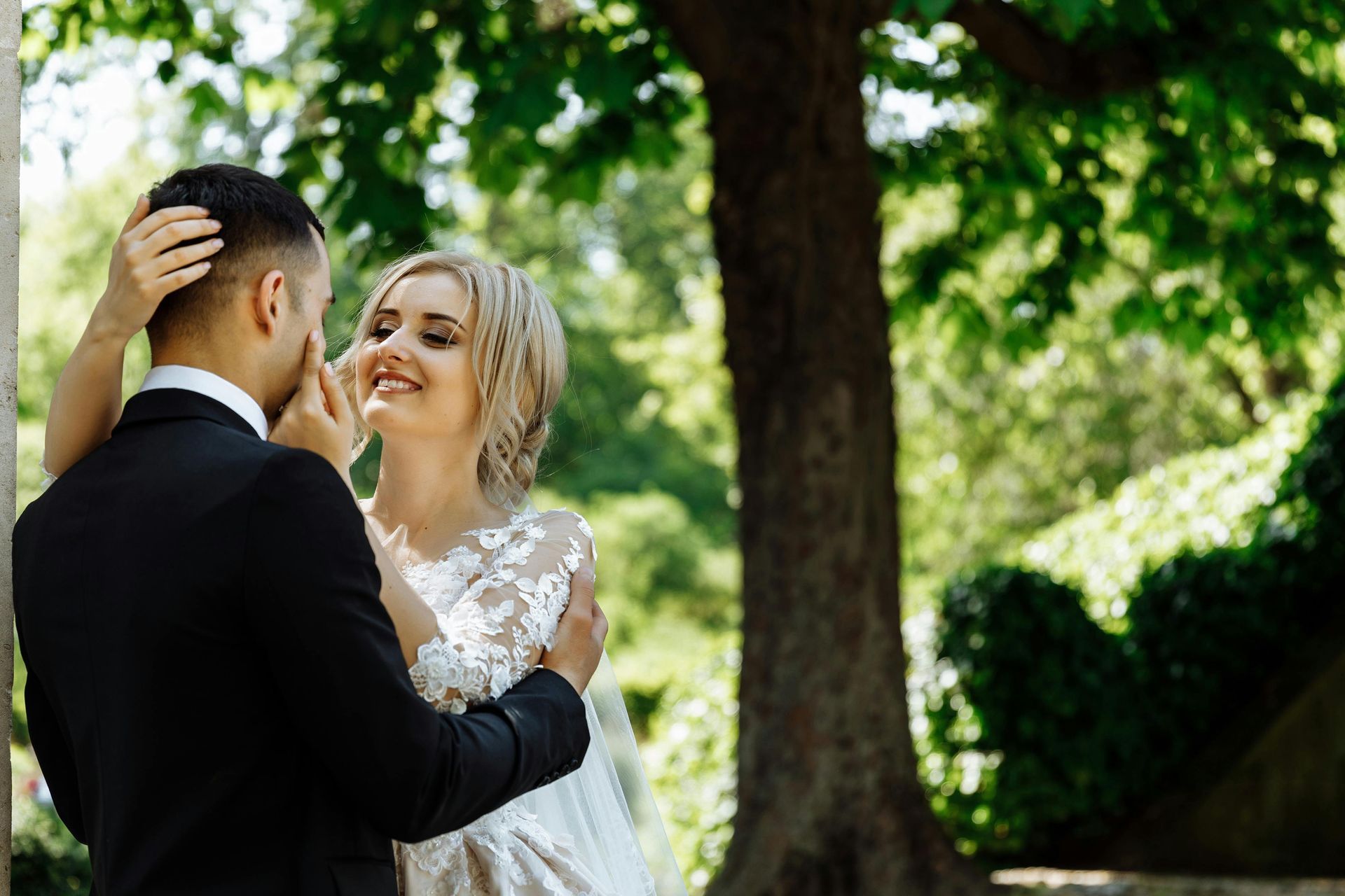 Bride and groom embrace outdoors; she smiles at him while he gently touches her face.