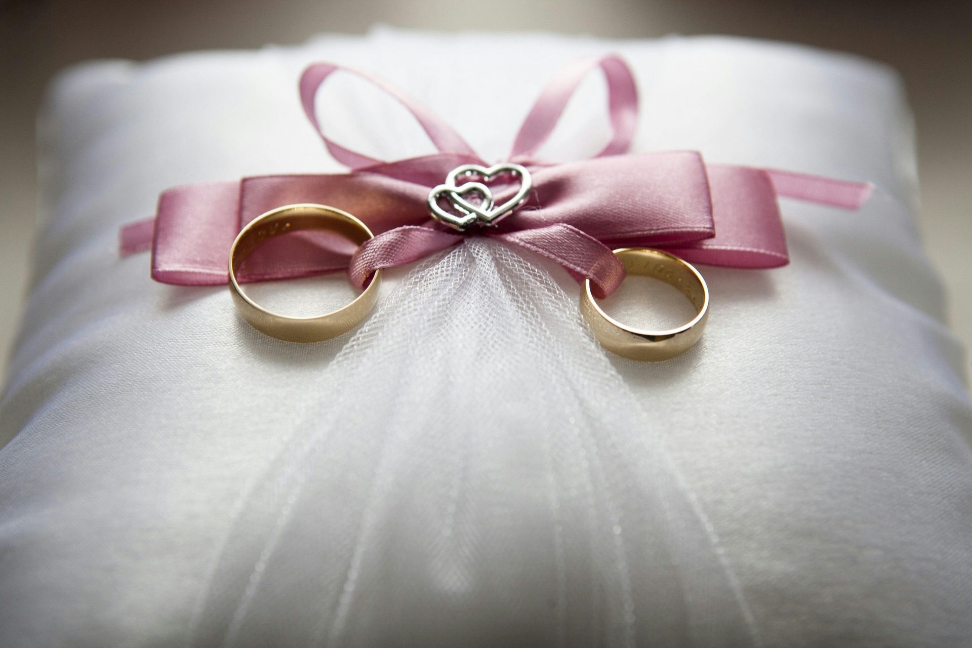Two gold wedding rings on a white pillow, adorned with a pink bow and a small silver heart.