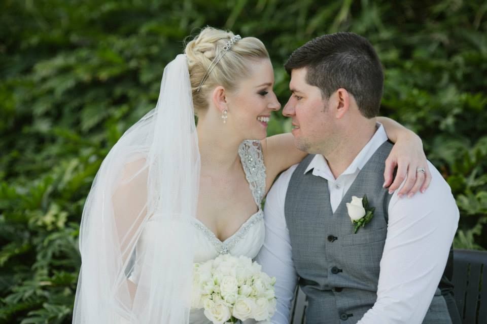 Bride and groom smiling at each other, outdoors. Bride in a wedding dress, veil and flowers, groom in a vest.