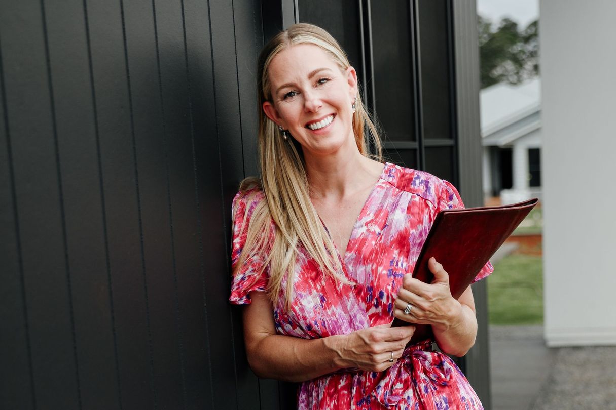 Blonde woman smiles, holding a red folder, leaning against a black wall.