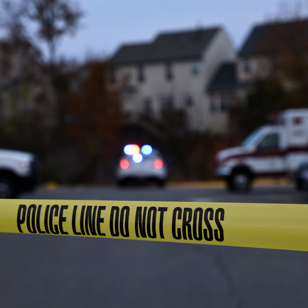 Police tape at a crime scene with a police car and ambulance in the background.