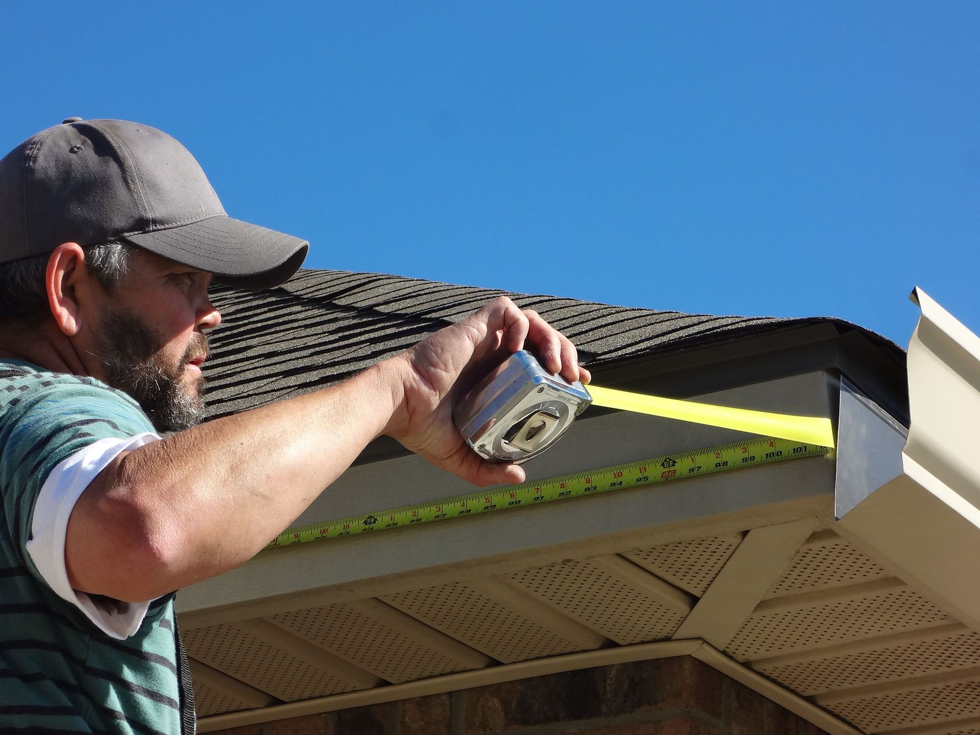 Man measures a gutter with a yellow tape measure under a blue sky.