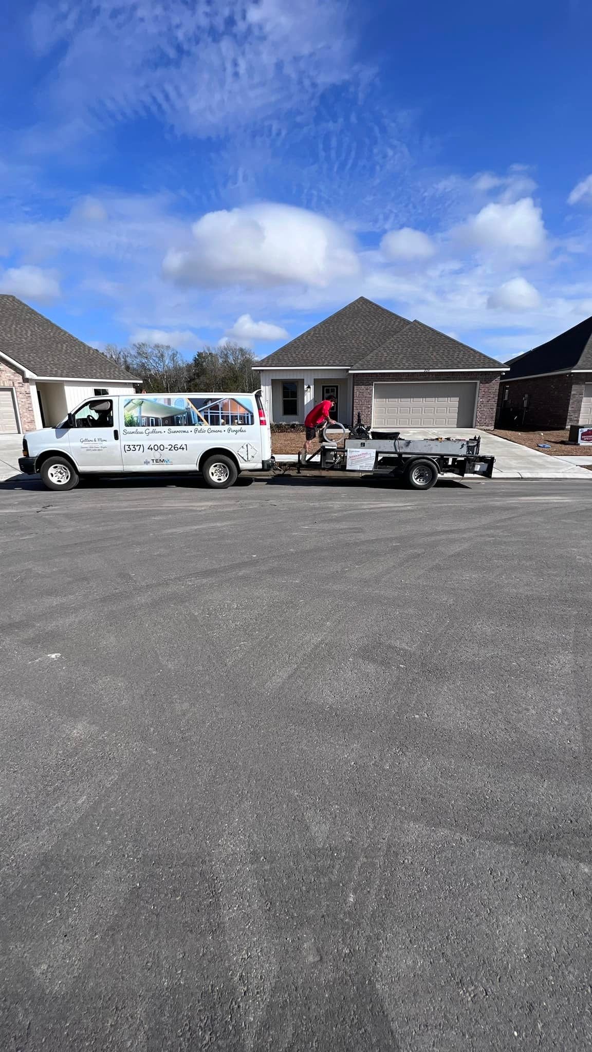 White van towing a trailer in front of new houses. Person in red jacket. Blue sky.