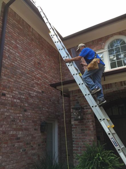 Man on a tall ladder cleaning gutters of a brick house. He wears jeans, a blue shirt, and a tool belt.