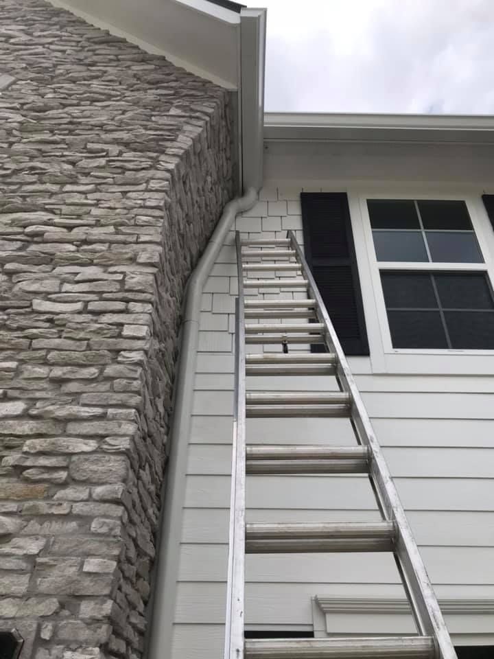 Ladder leaning against a house, near a stone chimney and white siding, for gutter work.